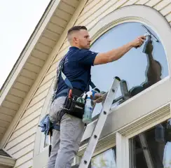 Professional window cleaner on a ladder using a squeegee to clean an exterior arched window, Stockton, CA.